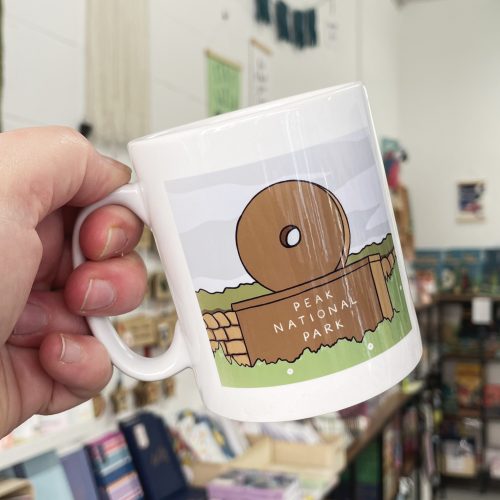 Peak District Millstone Mug An illustration of a Peak District National Park sign on a Millstone. The illustration is printed onto a white mug which is being held by the handle.