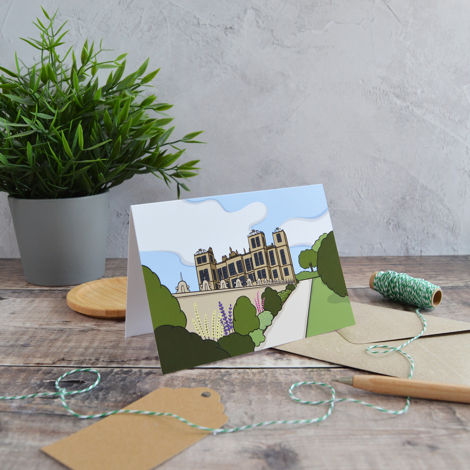An A6 landscape greetings card of Hardwick Hall. It is stood on the table with a kraft envelope, alongside a gift tag, wooden pen, green twine and a plant.