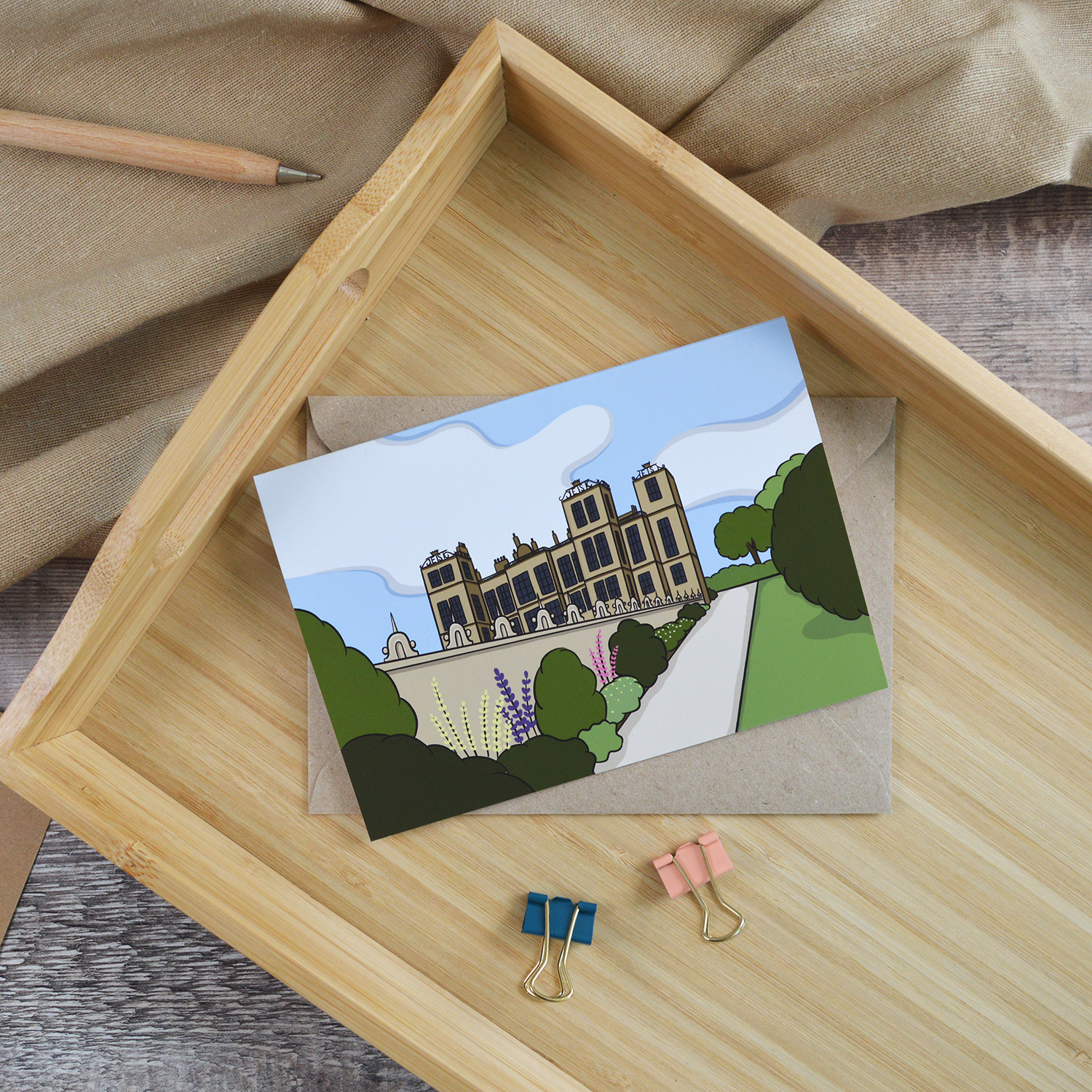 An A6 landscape greetings card of Hardwick Hall. It is laid down on a wooden tray alongside a wooden pen and two coloured bulldog clips
