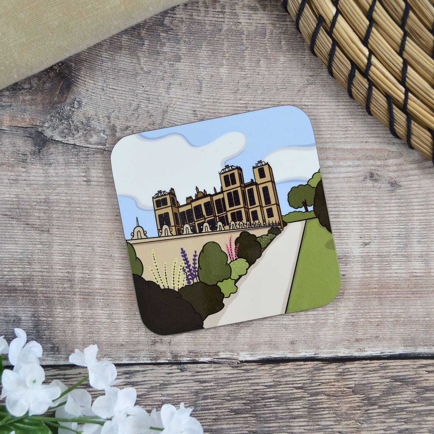 A coaster with an illustration of Hardwick Hall sat on a wooden table with white flowers and a natural placemat