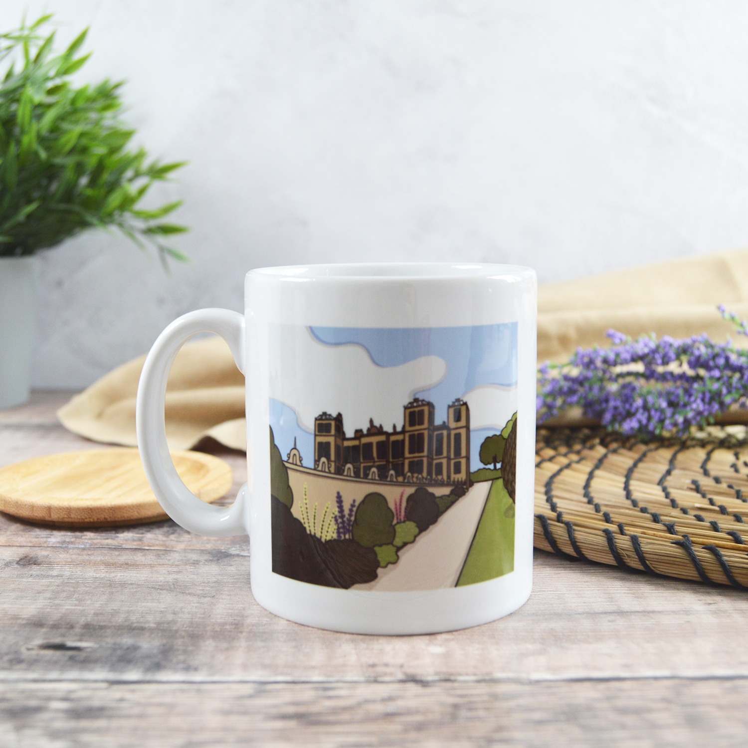 White mug printed with an illustration of Hardwick Hall. The mug is sat on a wooden table in front of a natural placemat and plant.