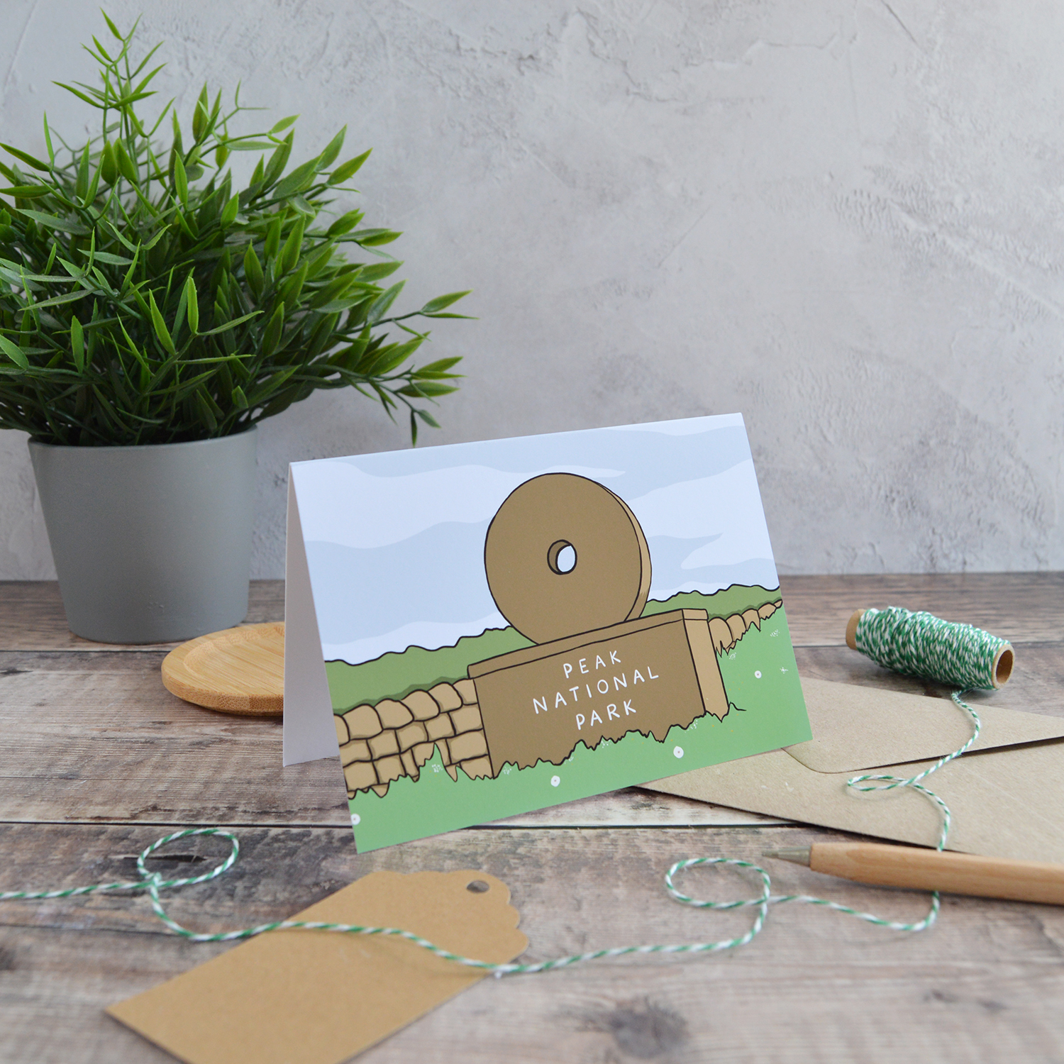 An A6 landscape greetings card of a Peak District Millstone Signs. It is stood on the table with a kraft envelope, alongside a gift tag, wooden pen, green twine and a plant.