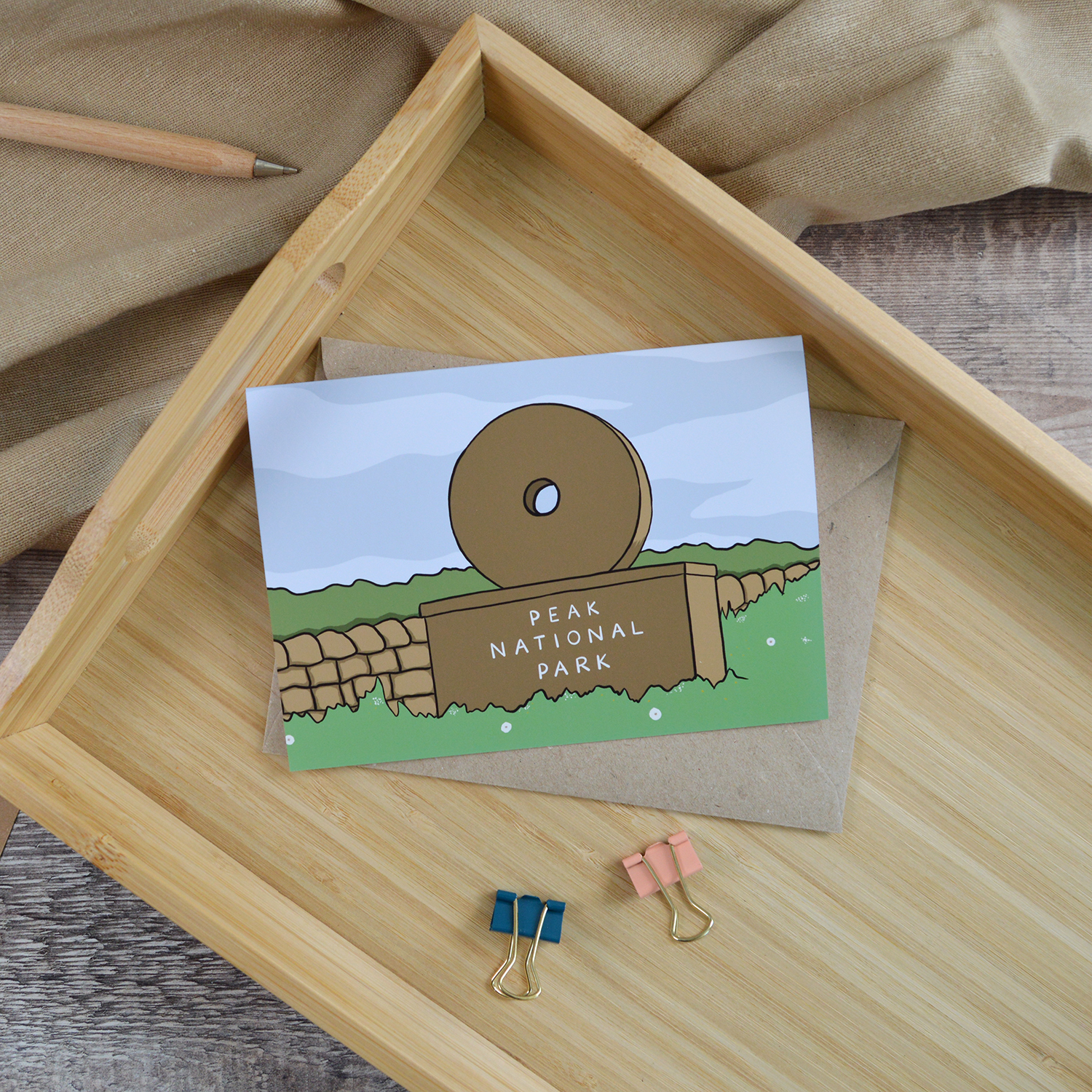An A6 landscape greetings card of a Peak District Millstone Sign. It is laid down on a wooden tray alongside a wooden pen and two coloured bulldog clips