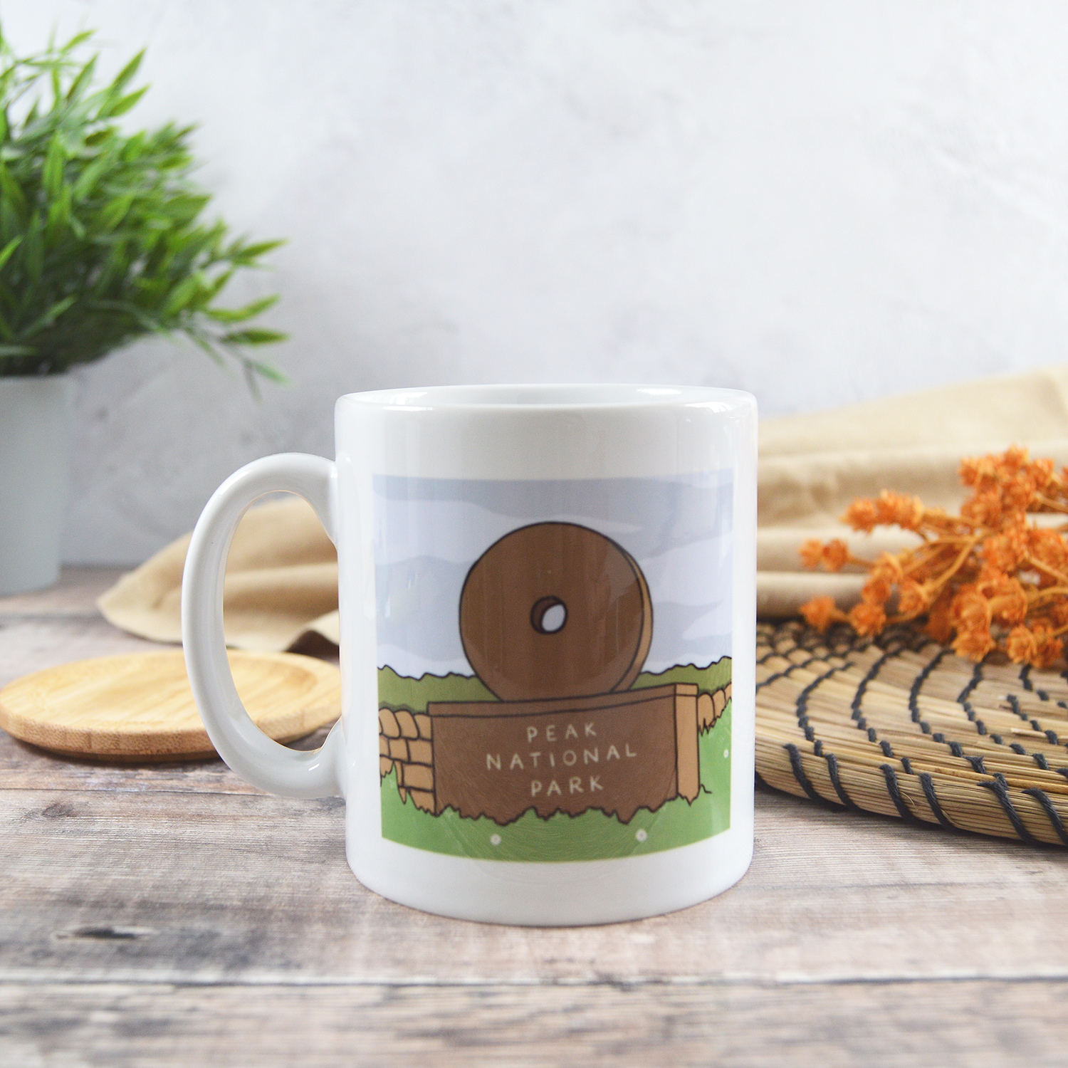 White mug printed with an illustration of a Peak National Park Millstone Sign. The mug is sat on a wooden table in front of a natural placemat and plant.