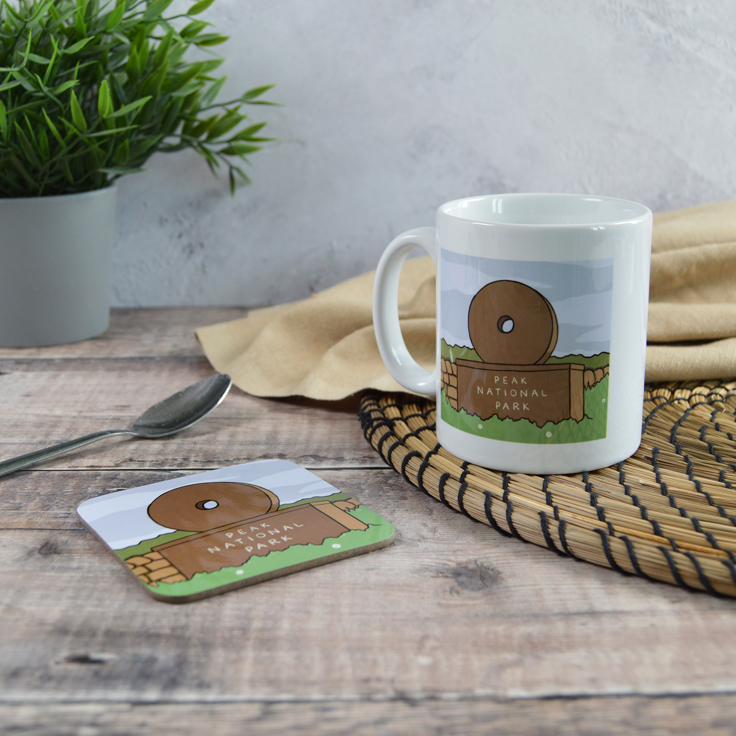 A white mug and a coaster with matching illustrations of The Peak District Millstone Sign. They are sat on a wooden table with a natural placemat, cloth, silver teaspoon and a plant.