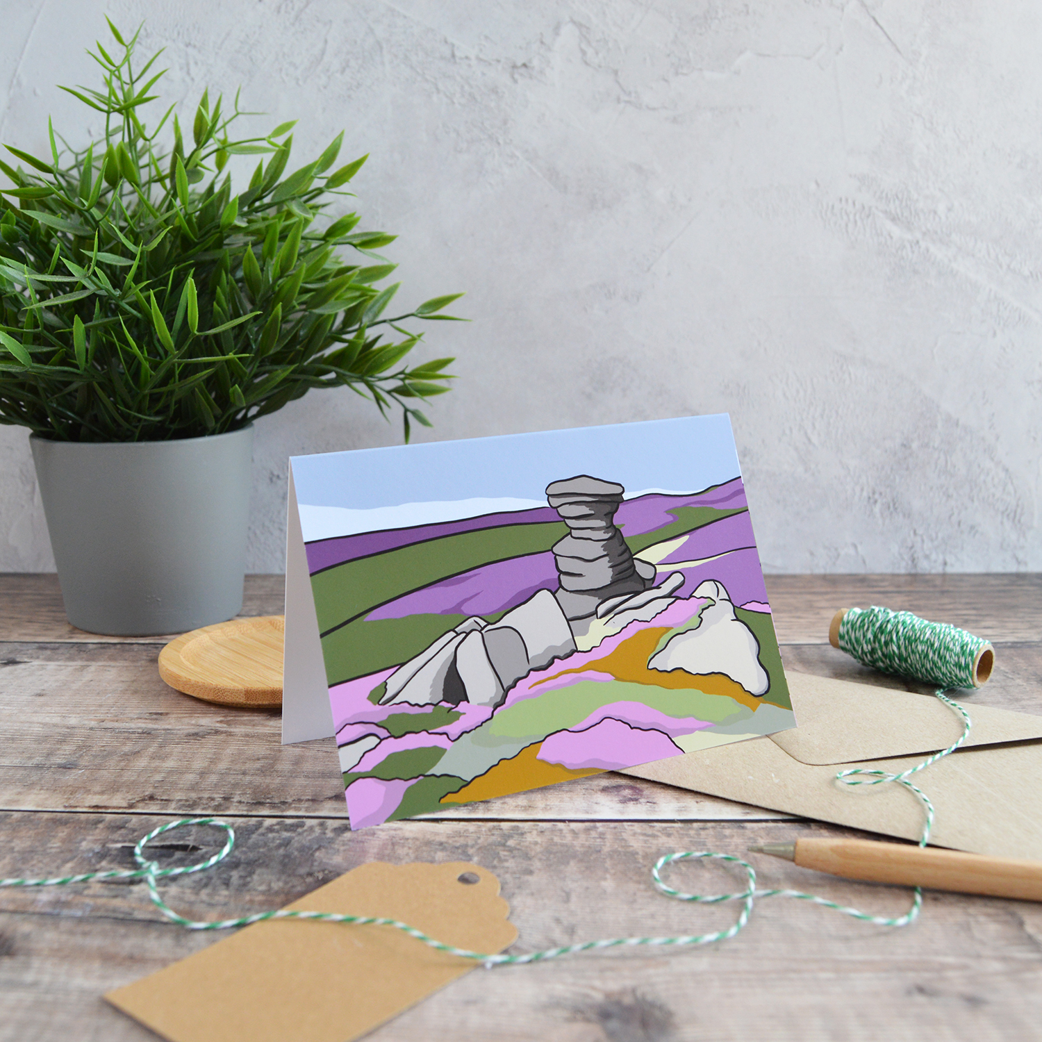 An A6 landscape greetings card of The Salt Cellar at Derwent Edge. It is stood on the table with a kraft envelope, alongside a gift tag, wooden pen, green twine and a plant.