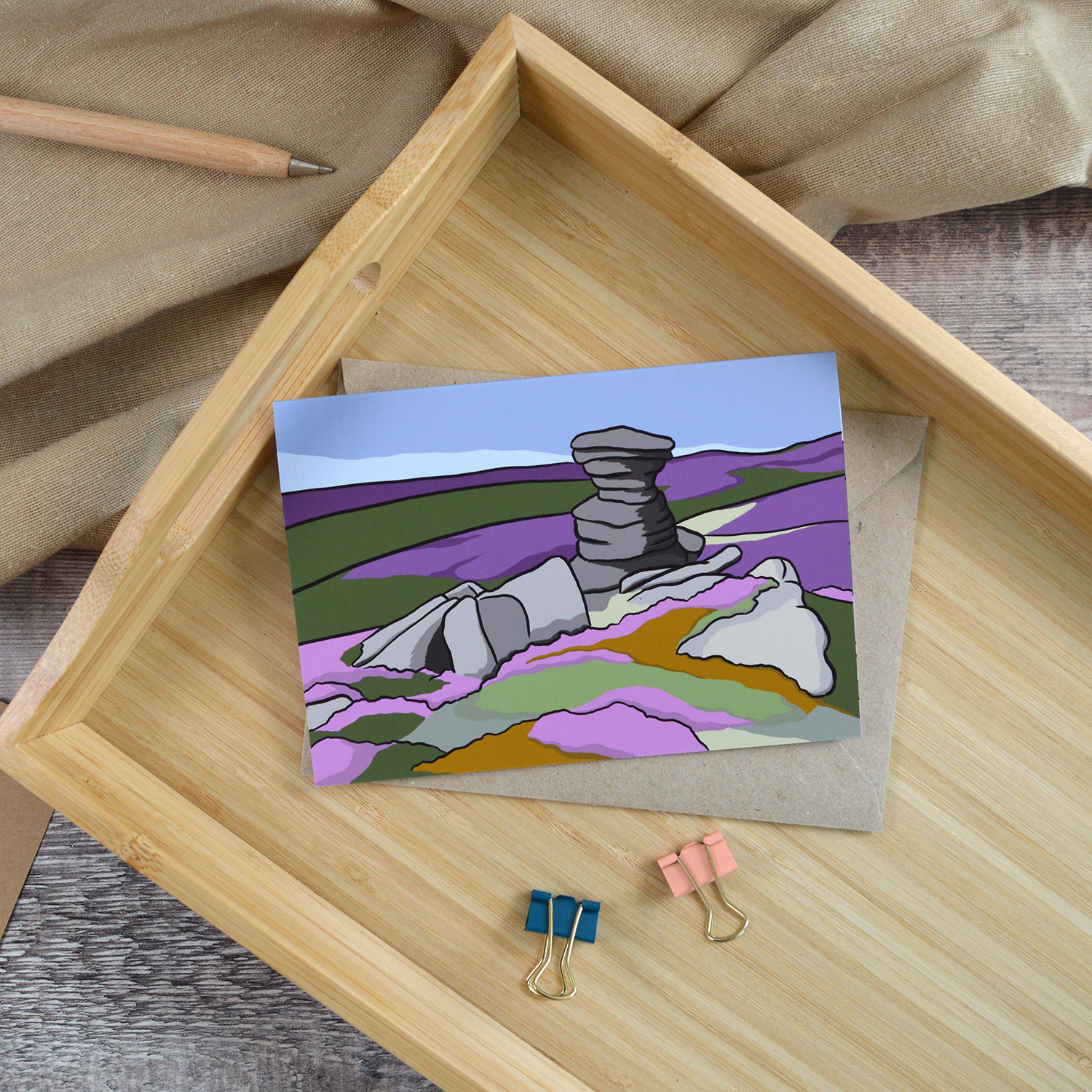 An A6 landscape greetings card of The Salt Cellar at Derwent Edge. It is laid down on a wooden tray alongside a wooden pen and two coloured bulldog clips