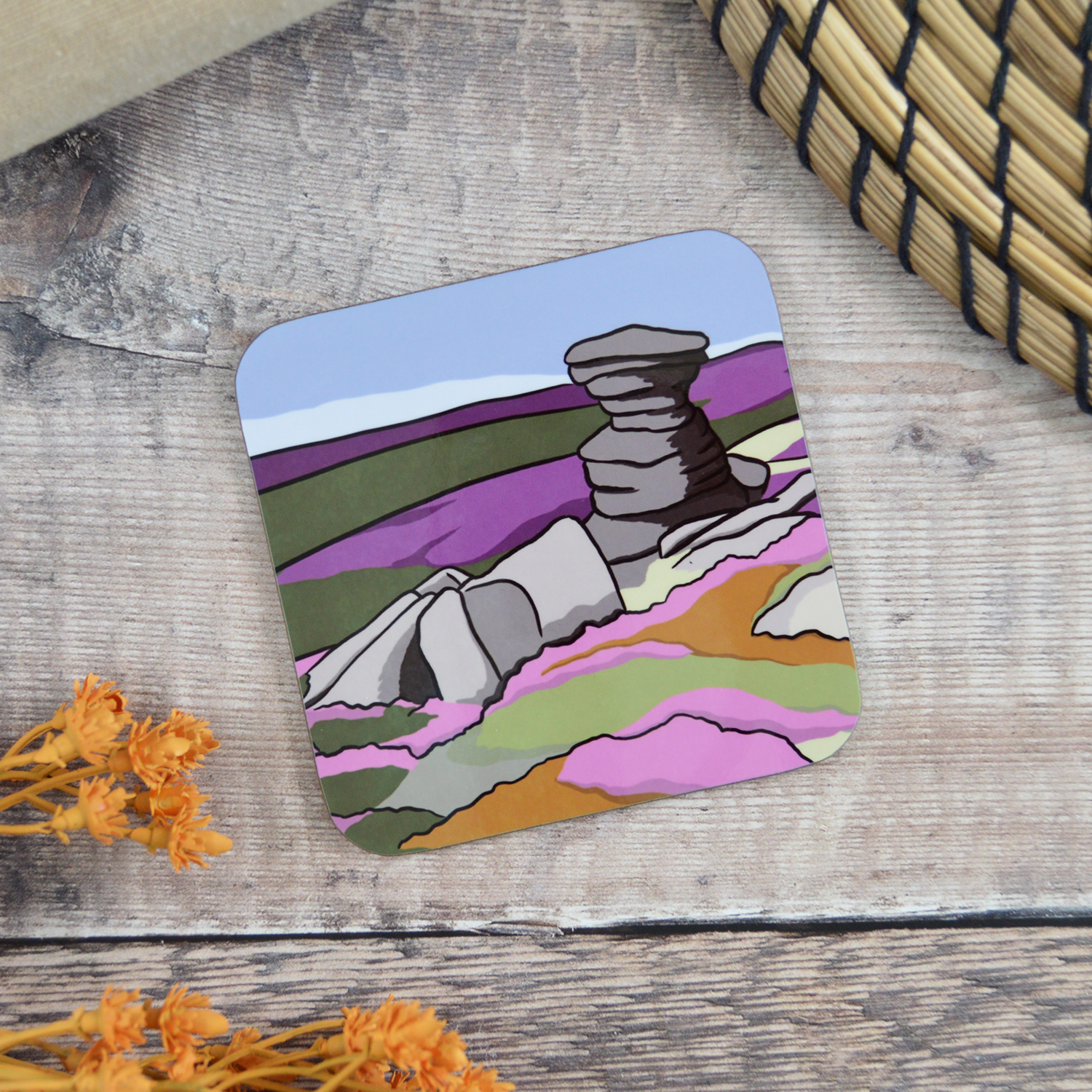 A coaster with an illustration of The Salt Cellar sat on a wooden table with orange flowers and a natural placemat