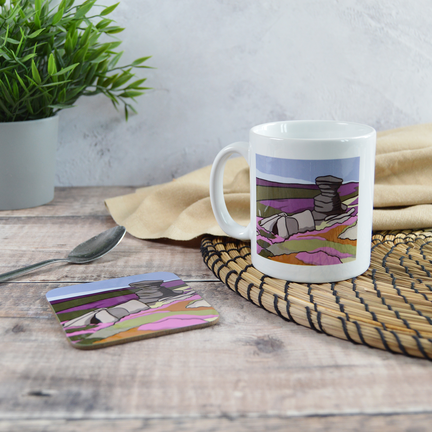 A white mug and a coaster with matching illustrations of The Salt Cellar. They are sat on a wooden table with a natural placemat, cloth, silver teaspoon and a plant.