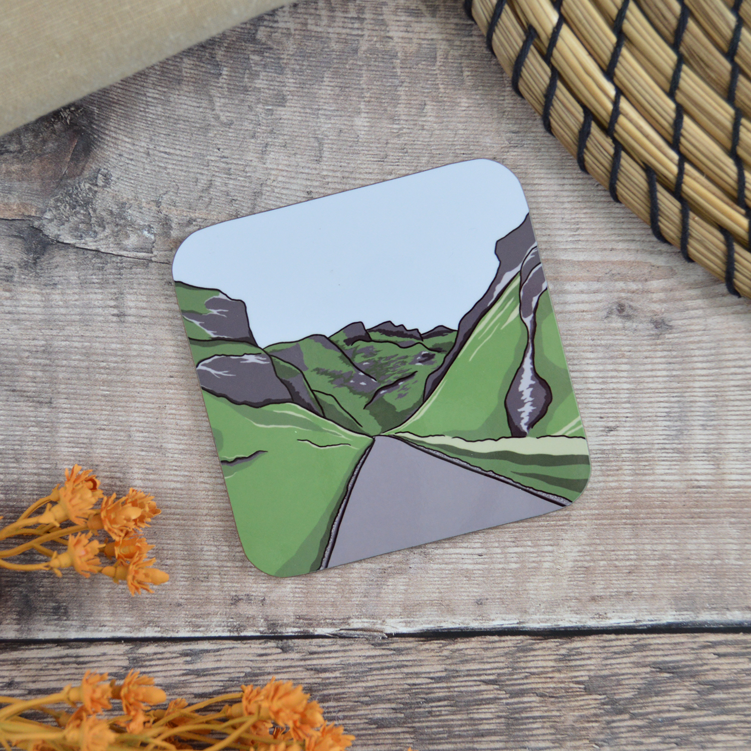 A coaster with an illustration of Winnats Pass sat on a wooden table with orange flowers and a natural placemat