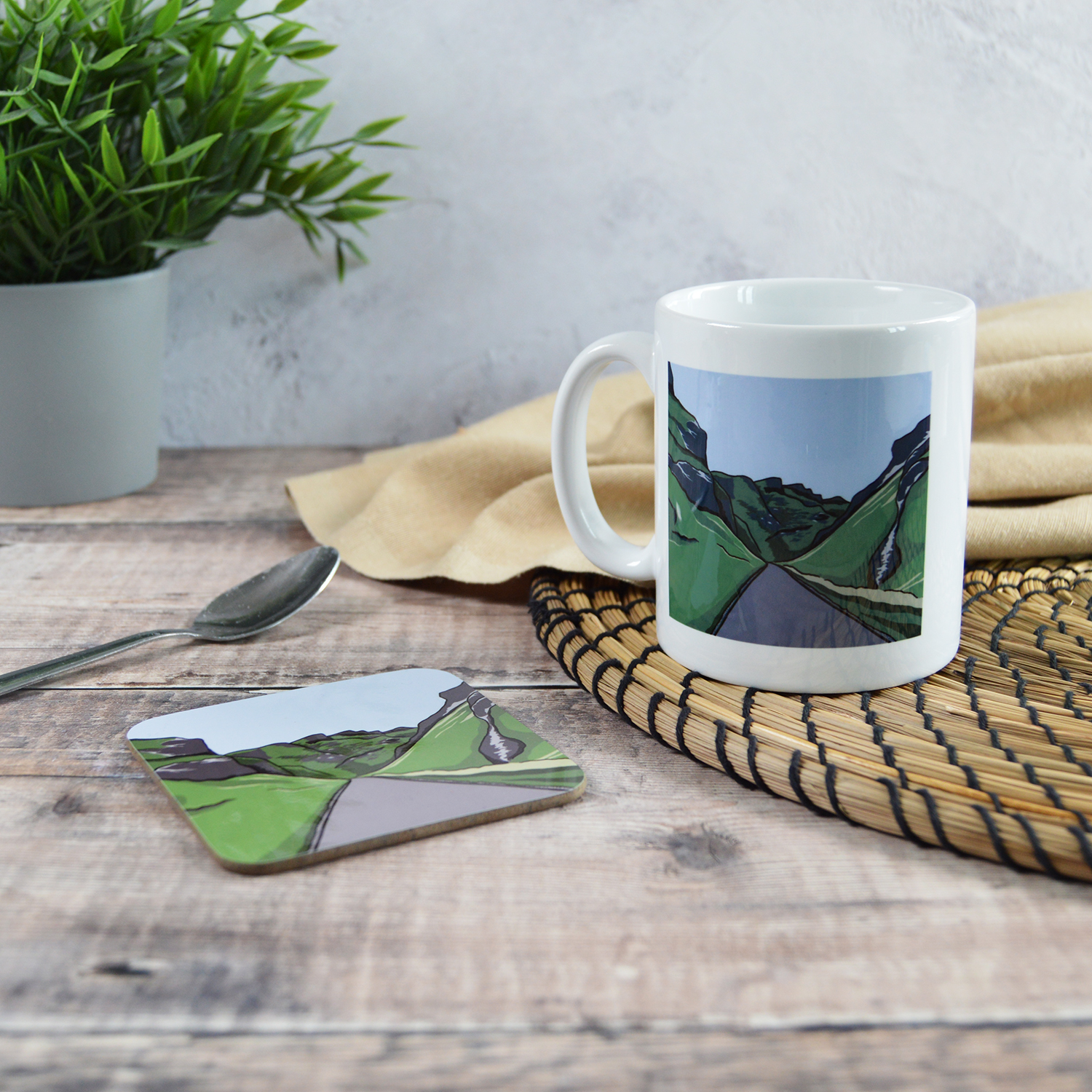 A white mug and a coaster with matching illustrations of Winnats Pass. They are sat on a wooden table with a natural placemat, cloth, silver teaspoon and a plant.